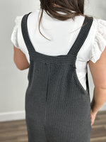 Load image into Gallery viewer, View of the shoulder straps on the Kobe Ribbed Comfort Jumpsuit in charcoal. Layered with a white flutter sleeve women&#39;s top.
