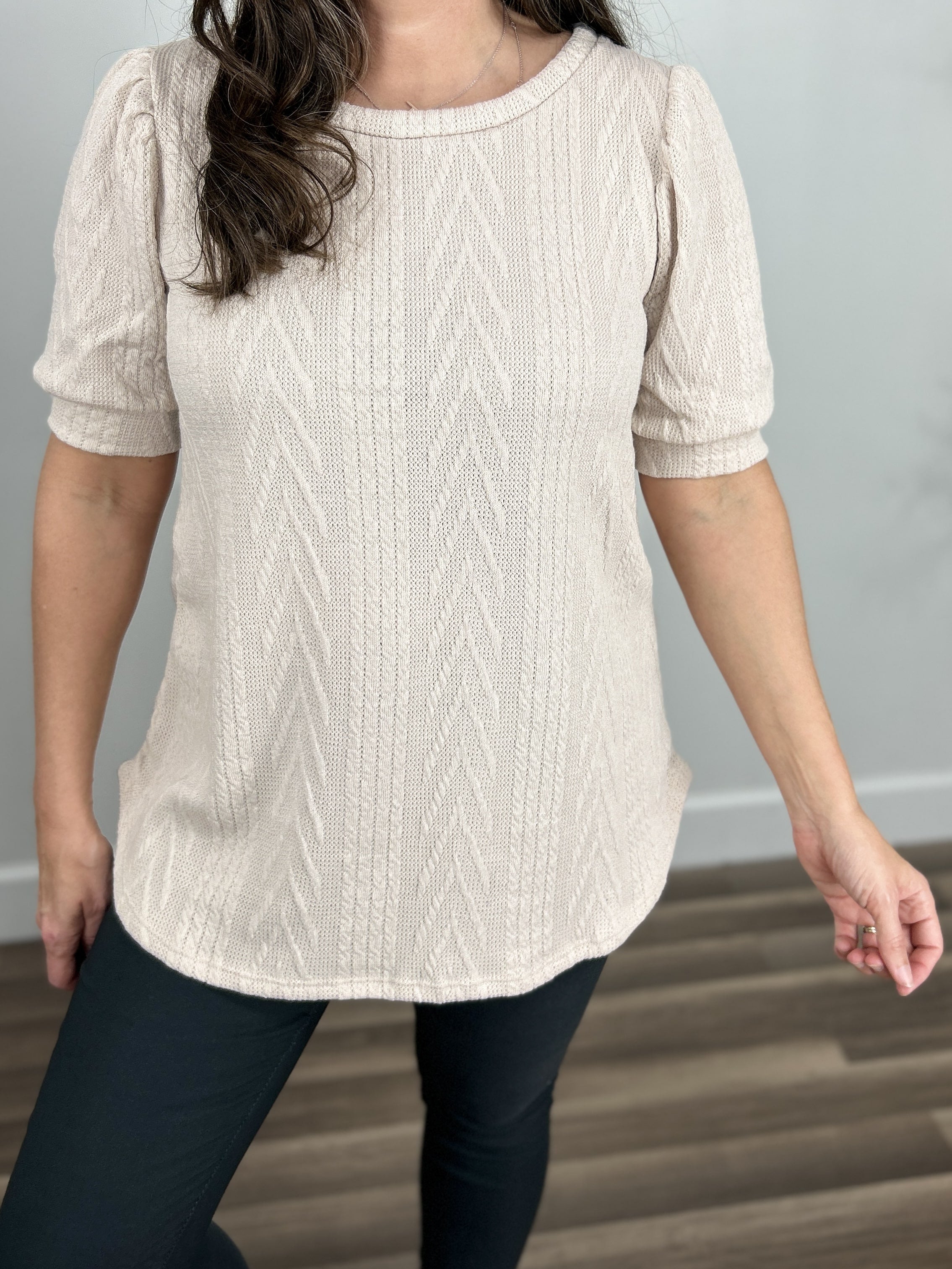 Detailed view of the cable knit fabric of the oatmeal color lacy top with a round neckline and curved hemline.