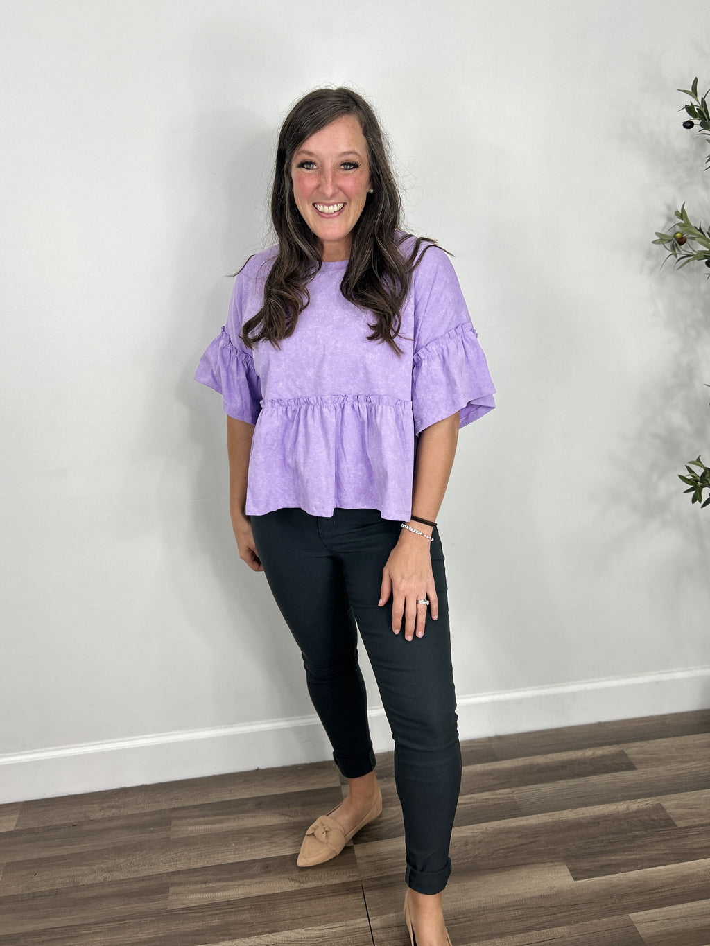 Women's purple peplum ruffle sleeve top paired with charcoal skinny jeans and camel color flat slip on shoes.
