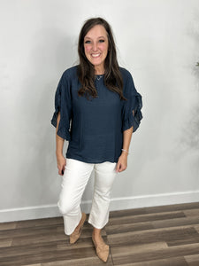 Women's navy three quarter ruffle sleeve top paired with white crop pants and camel flat shoes.