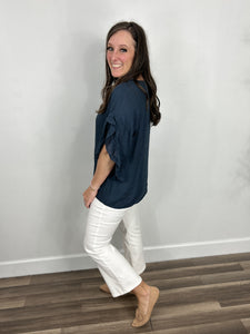 Side view of the Rigsby top in navy showing the ruffle three quarter sleeves and straight hemline. Paired with white crop pants and flat camel color shoes.