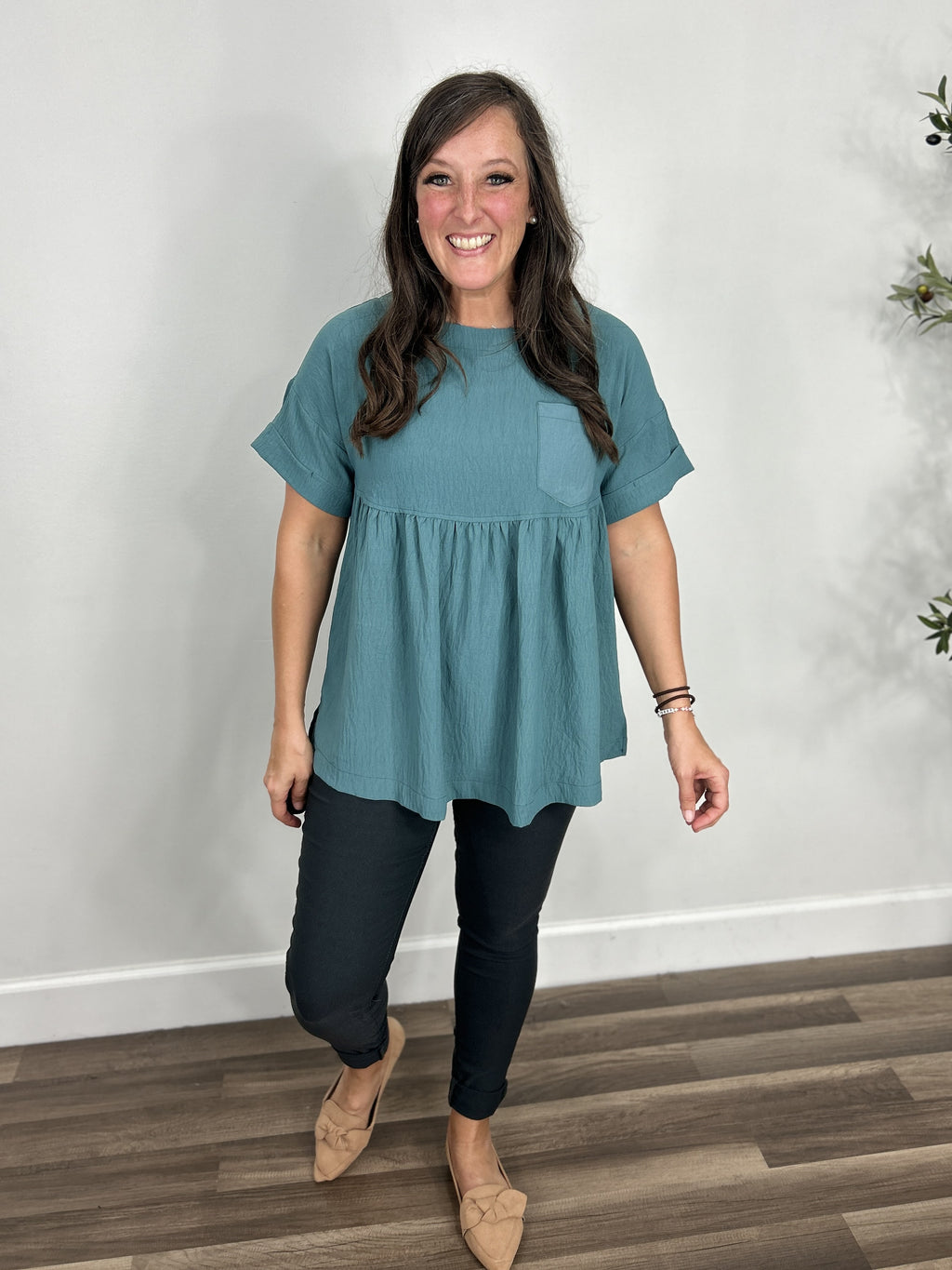 Women's teal short sleeve babydoll top paired with charcoal skinny jeans and camel color slip on shoes.